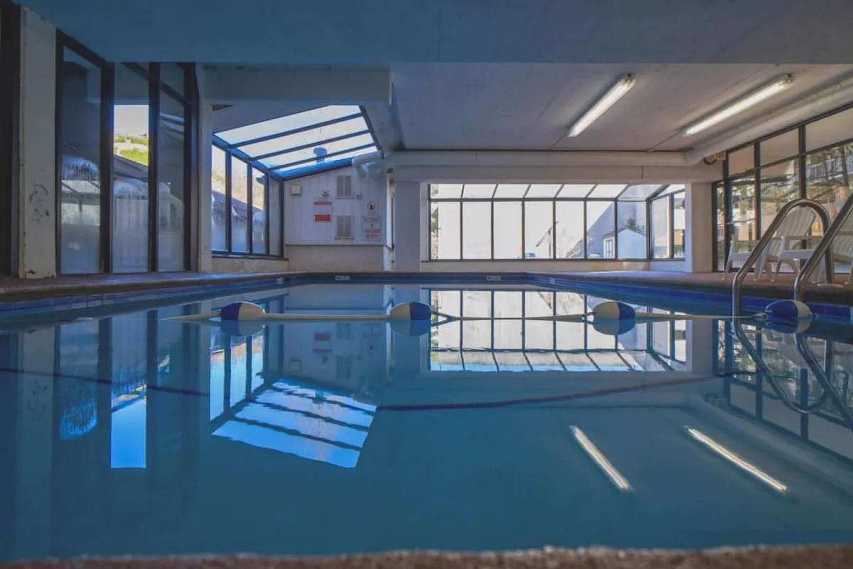 An interior shot of the swimming pool area, with sunlight streaming through the glass roof, children’s pool toys floating, and the pool flanked by windows and steps. The atmosphere signals fun and safe recreation for families, especially kids.