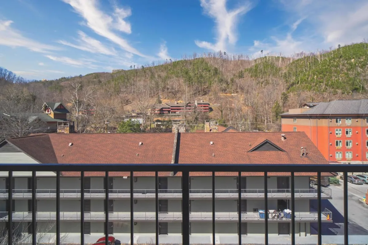 A tranquil balcony scene overlooking Gatlinburg's skyline and the surrounding mountain landscape. The mountain view is framed by a classic balcony railing, while the red-roofed buildings below add a charming local touch. The mood is quiet and romantic, suggesting a perfect spot for couples to relax and enjoy the breathtaking scenery together.