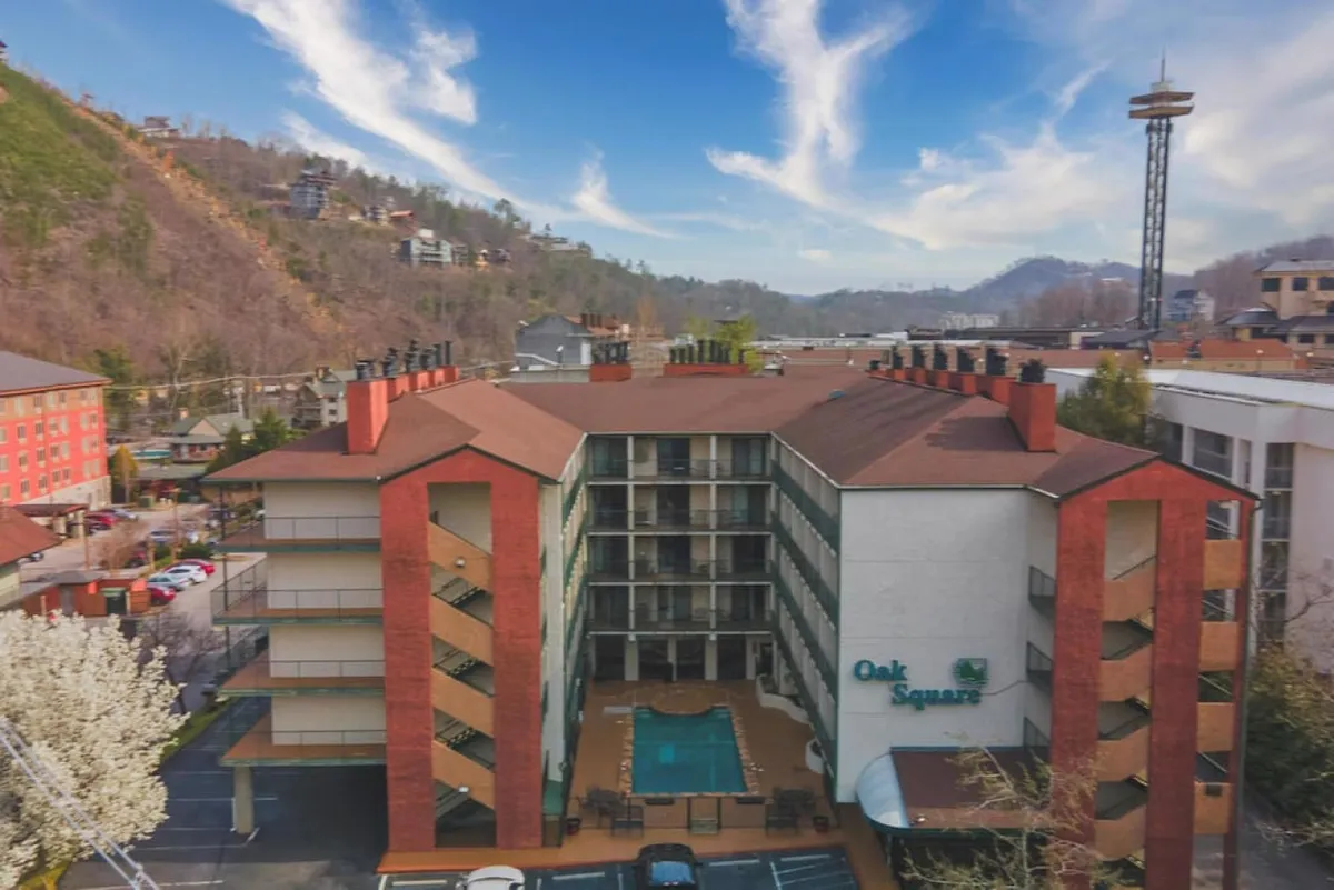 An exterior view of the hotel’s outdoor pool, positioned in front of the building, with the surrounding hills visible in the backdrop. Guests are sunbathing by the pool, swimming, and relaxing on deck chairs. The hotel facade and balconies are also visible, evoking a resort vibe.