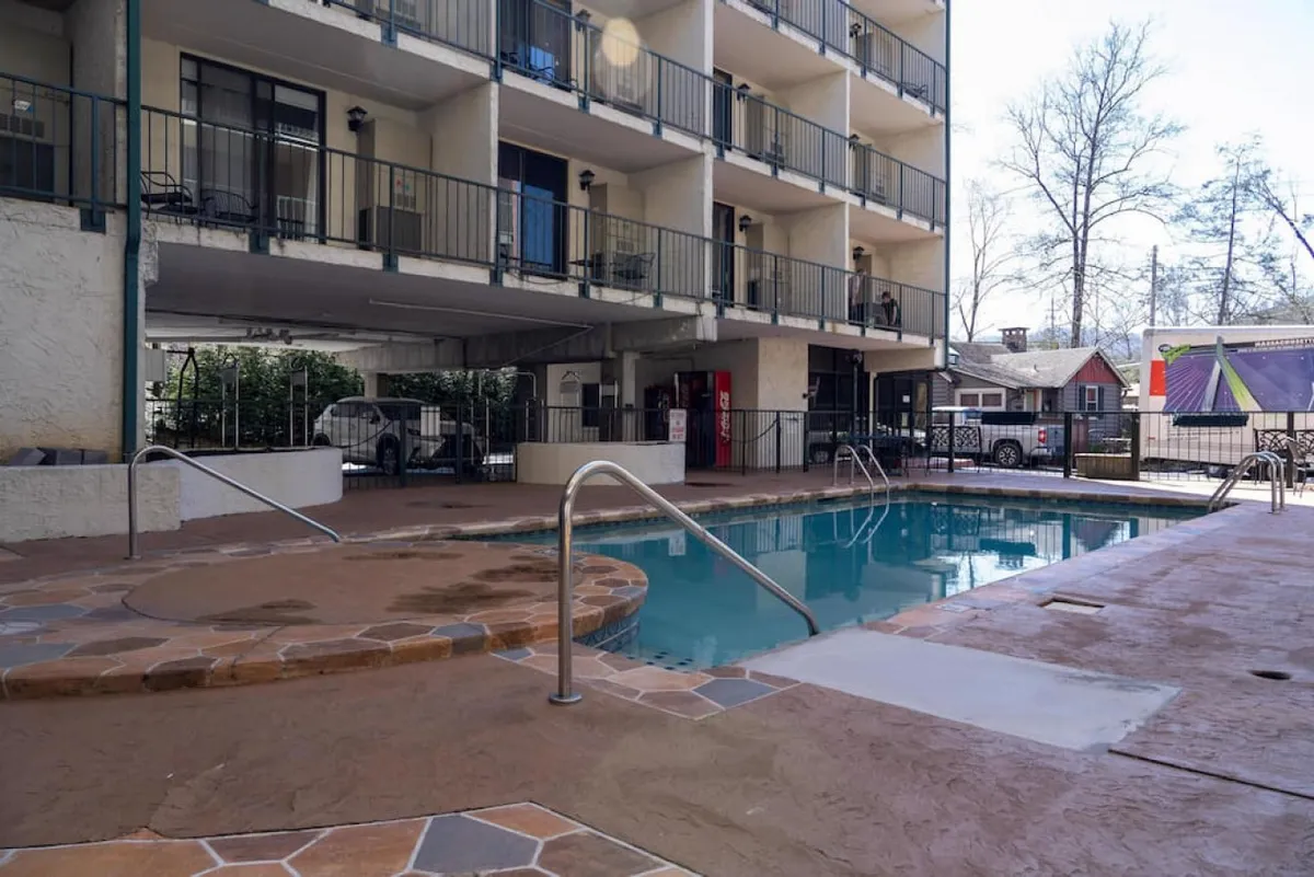 A cheerful scene at the outdoor pool area, with a hot tub visible beside the pool, guests relaxing on loungers, and balconies of the building in the background. The mood is relaxed and social, suggesting opportunities for sunbathing and poolside gatherings.