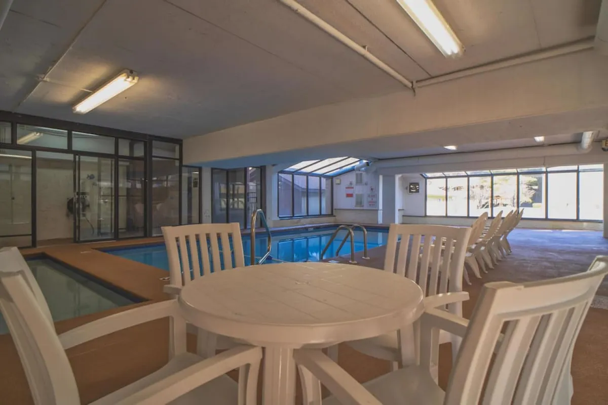 A spacious indoor pool area with sparkling blue water, white chairs, and a table for relaxing between swims. Natural light fills the room, creating a leisurely vibe that invites guests to unwind and swim together, regardless of the weather outside.
