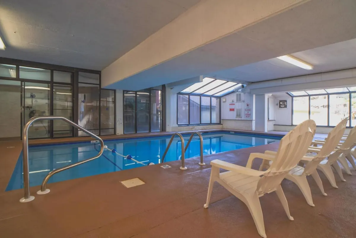A spacious indoor swimming pool bathed in sunlight streaming through skylights, with white lounge chairs arranged poolside. Several guests—families and friends—are swimming, relaxing, and socializing in a bright, inviting atmosphere. The pool’s shallow section is visible, ideal for children.